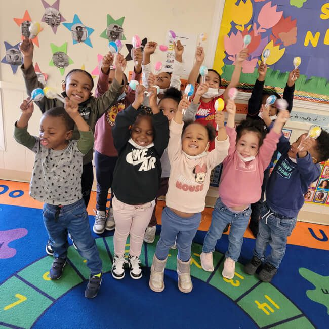 A group of children in a daycare hold up maracas in their hands and smile.