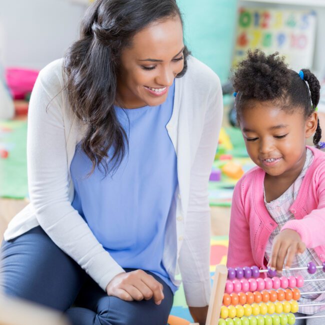 Mid adult female African American preschool teacher helps a preschool student with math. The little girl is using an abacus.