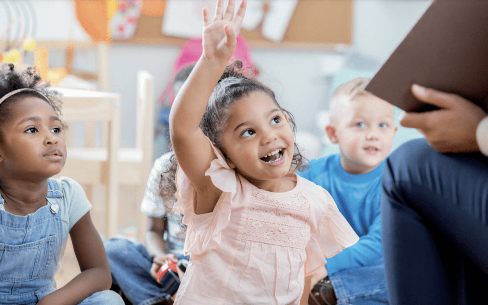 Young girl sitting in a classroom raising her hand with other students sitting around her