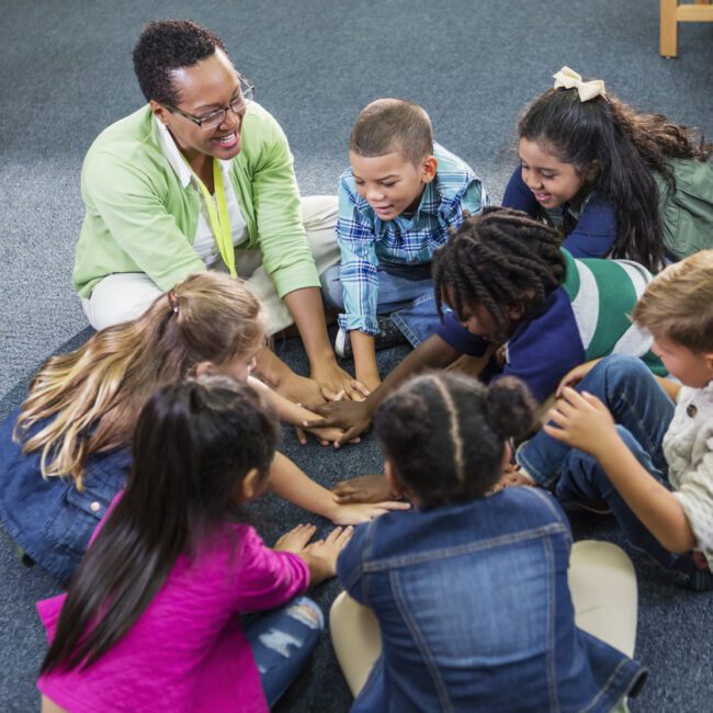 A female African-American teacher sitting on the floor in a circle with her elementary school class, a group of seven multi-ethnic children. They are playing a game, reaching, putting their hands in the middle of the circle.