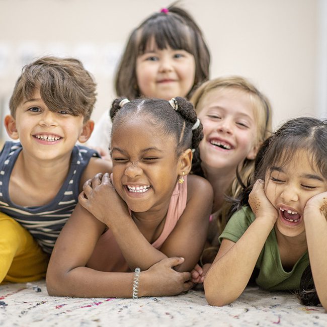 Multi ethnic group of preschool children all huddled together laying on the floor of their classroom as they pose for the camera. They are all happy and smiling.