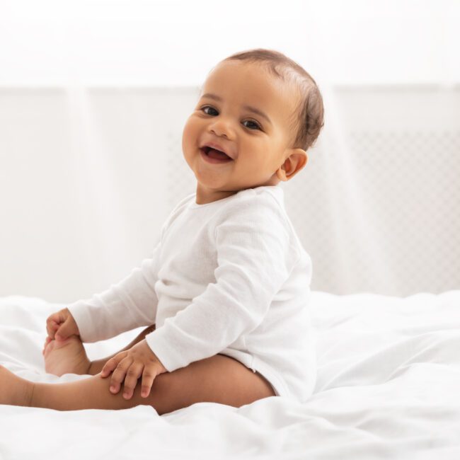 Portrait Of Adorable Baby Sitting On Bed And Smiling Looking At Camera