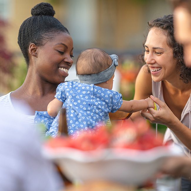 A young Black mother holds her young child in her lap at an outdoor dining table. A friend is smiling and reaching the baby