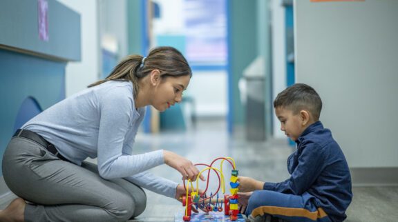 A young female occupational therapist is seated across a young male patient. They are both focused on playing with a bead maze that is in between them. They are both dressed casually for their appointment together.