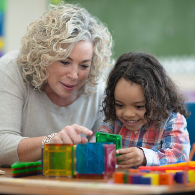 A boy of African descent sits at a table in his classroom one afternoon. Him and his teacher are building a cube together out of magnetic plastic building pieces. They are focused but also smiling and having fun.