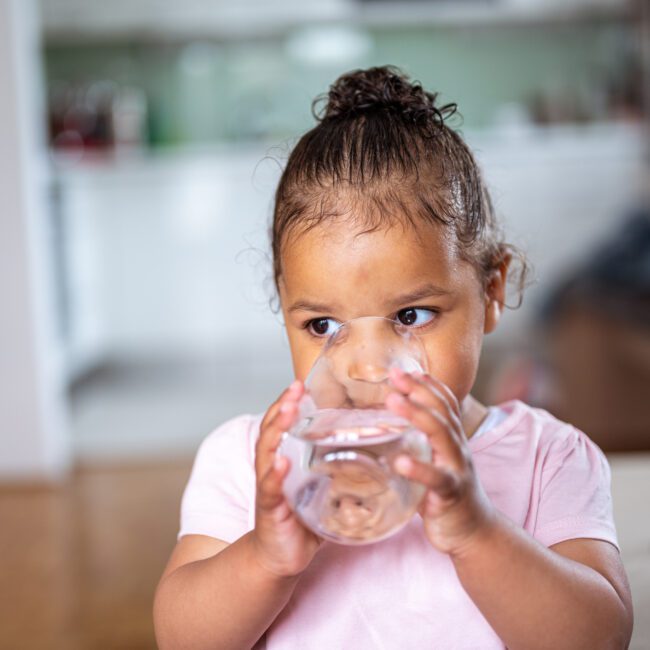Little cute african american girl drinking water from the glass in a domestic room.