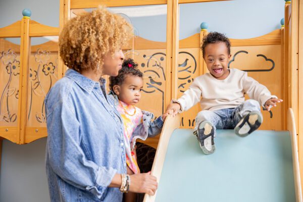 A young child happily slides down at an indoor playground while another child and child care provider look on.