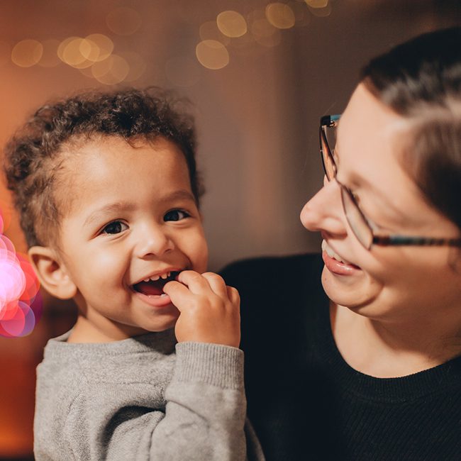 Child with finger near mouth held by a smiling adult with glasses and a black top. Dappled light behind them.